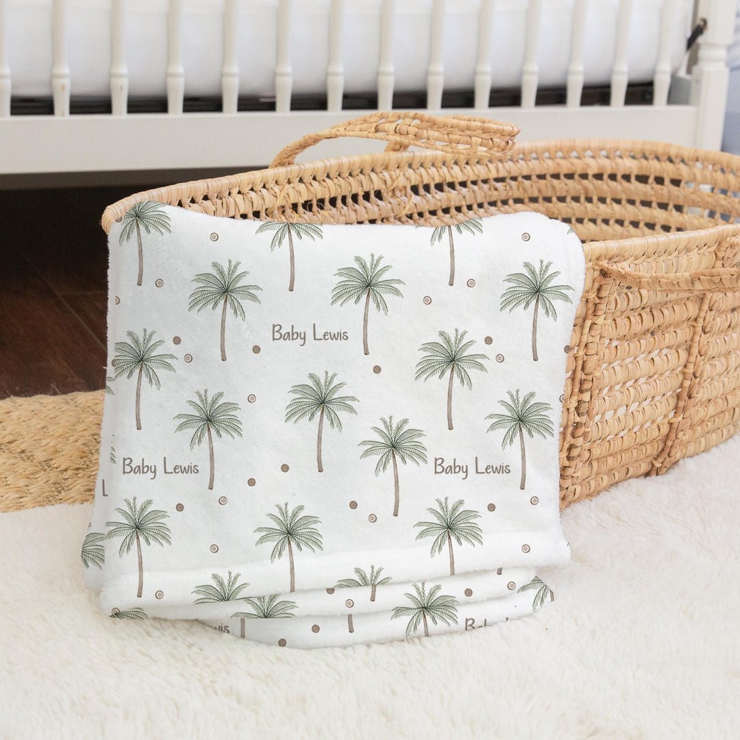 Baby blanket with palm tree pattern on a white surface next to a wicker basket.